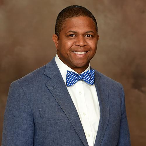 Mr. Donovan Lee-Sin, an African American male, smiles at the camera wearing a light blue suit with a white collared shirt and blue and white striped bow tie.