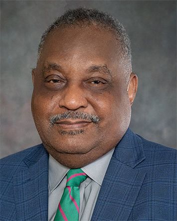 Mr. James Kendrick, an African American male with salt and pepper hair and a goatee, grins at the camera wearing a blue plaid suit with a grey collared shirt and a mint and fushcia striped tie.