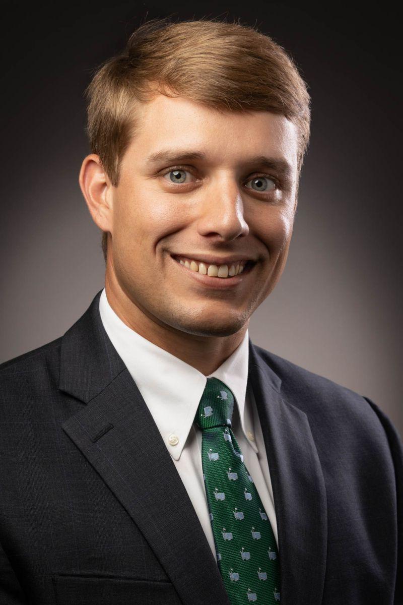 Mr. Nicholas Wood, a Caucasian male, smiles wearing a black suit with a white button down shirt and a green tie decorated with white apple symbols.